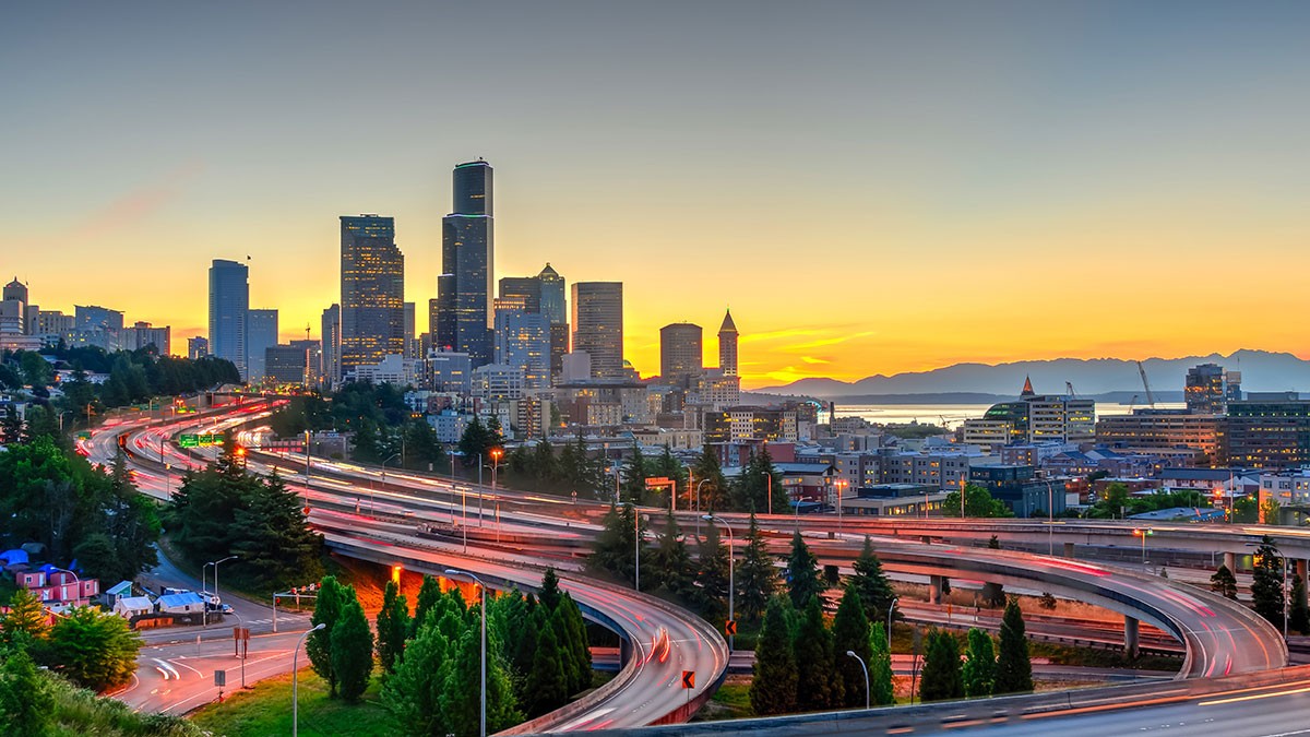 A vibrant cityscape at sunset with a skyline of tall buildings, winding highways with light trails, and a golden sky over distant mountains.