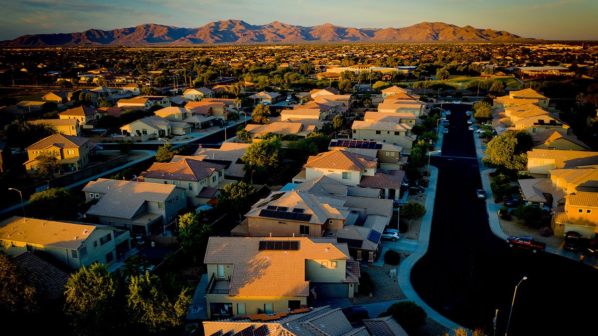 Aerial view of a suburban neighborhood at sunset, with rows of houses, tree-lined streets, and mountains in the background.