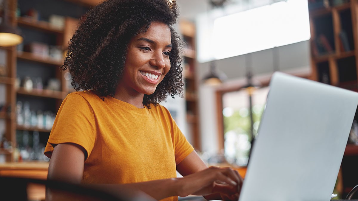 Smiling woman in a yellow shirt using a laptop in a cozy library or cafe setting with bookshelves in the background.