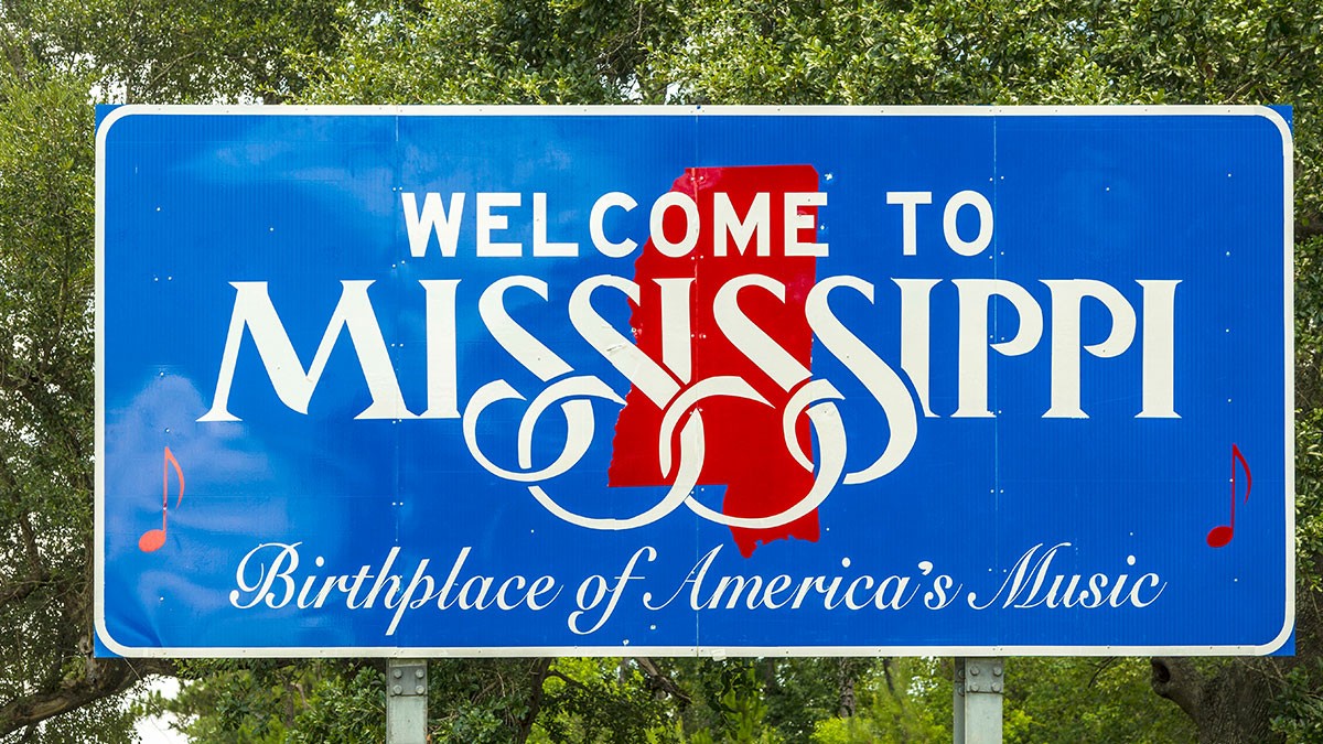 Blue sign with white text reading "Welcome to Mississippi, Birthplace of America's Music," featuring musical notes and a red state silhouette.