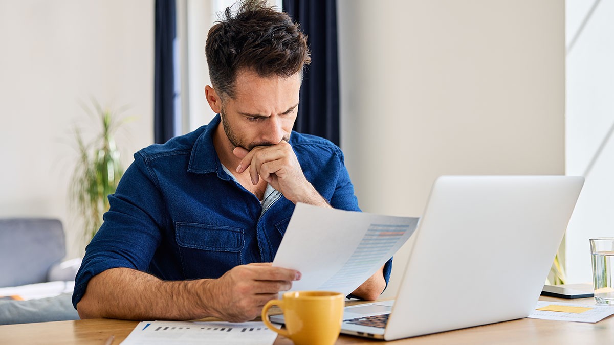 Man in a blue shirt sits at a desk, focused on reading a document, with a laptop and yellow mug nearby.