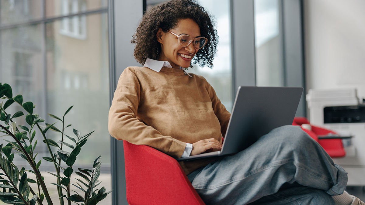 A person with curly hair and glasses smiles while using a laptop, sitting in a red chair next to a plant in a modern office setting.