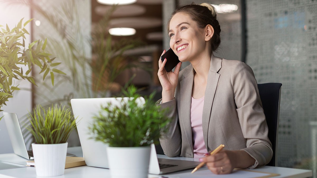 Smiling woman in a gray blazer talks on a phone at a desk with a laptop, surrounded by green plants.