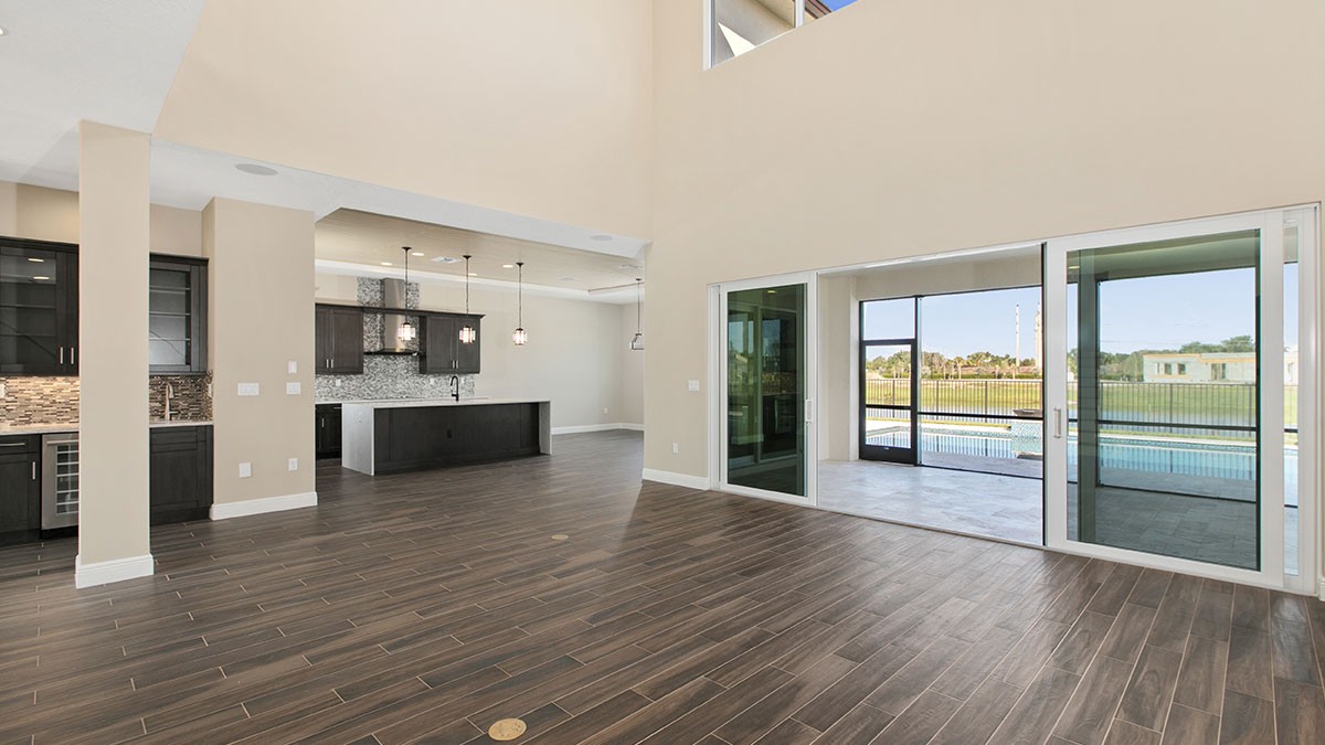 Spacious living area with wood flooring, open kitchen, and large glass doors leading to a patio with a view of a pool and greenery.