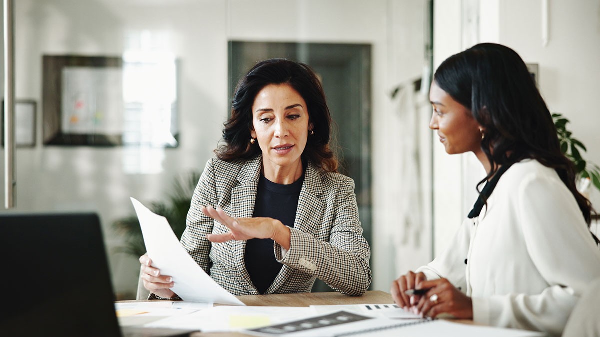 Two businesswomen in an office reviewing documents; one gestures while explaining a report to the other.