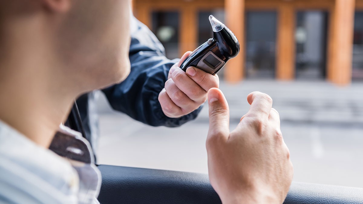 A person is holding a breathalyzer towards a driver seated in a car, suggesting a roadside sobriety test.
