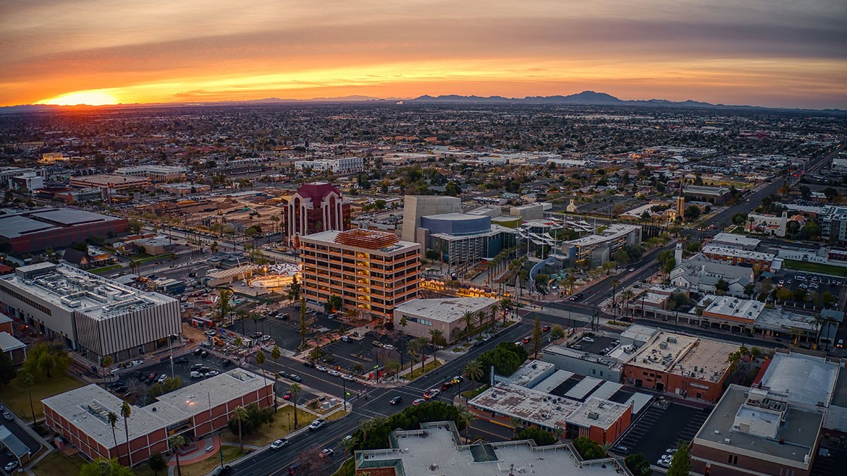 Aerial view of a city at sunset, with a mix of commercial and residential buildings, tree-lined streets, and distant mountains under a colorful sky.