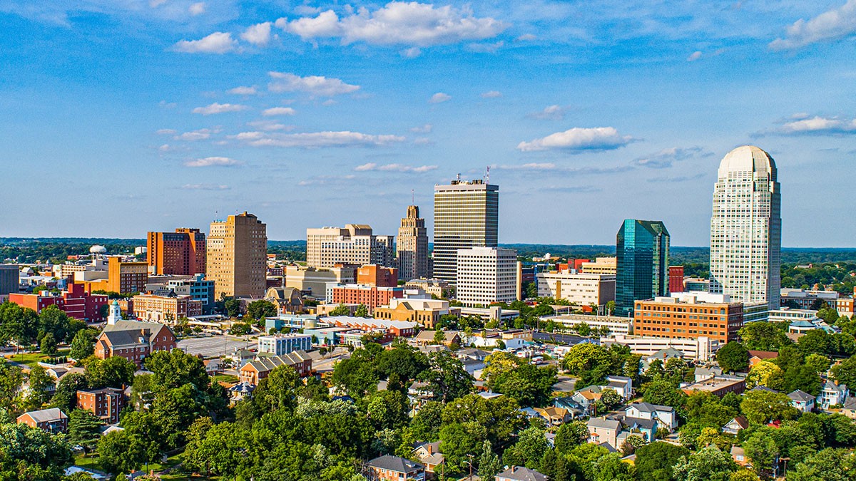 Aerial view of a mid-sized city's skyline with high-rise buildings, residential neighborhoods and green trees beneath a blue sky.