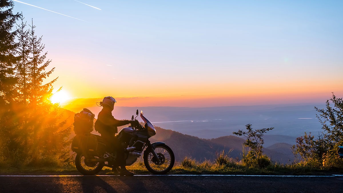 Silhouette of a motorcyclist riding along a road at sunset, with a scenic view of mountains and a glowing sky in the background.