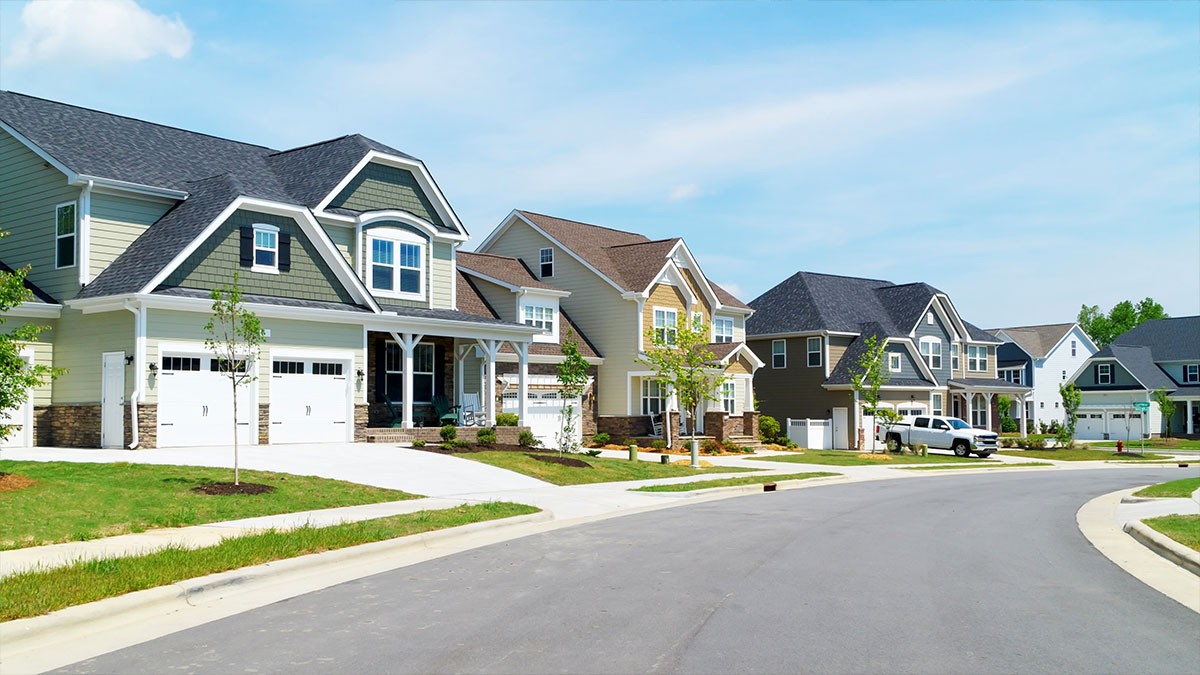 Suburban street with modern, two-story houses, neatly manicured lawns, and a clear blue sky. A few cars are parked in driveways.