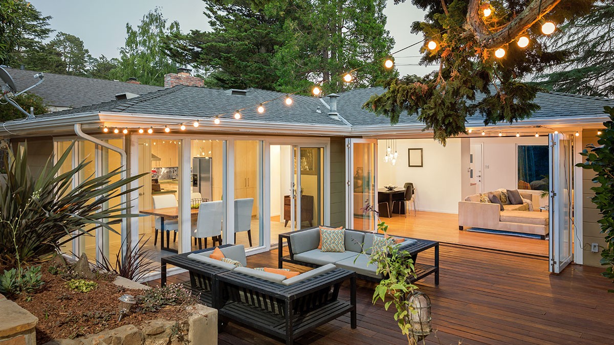 A cozy backyard patio with string lights, outdoor seating, and open sliding doors leading to a warmly lit living and dining area.