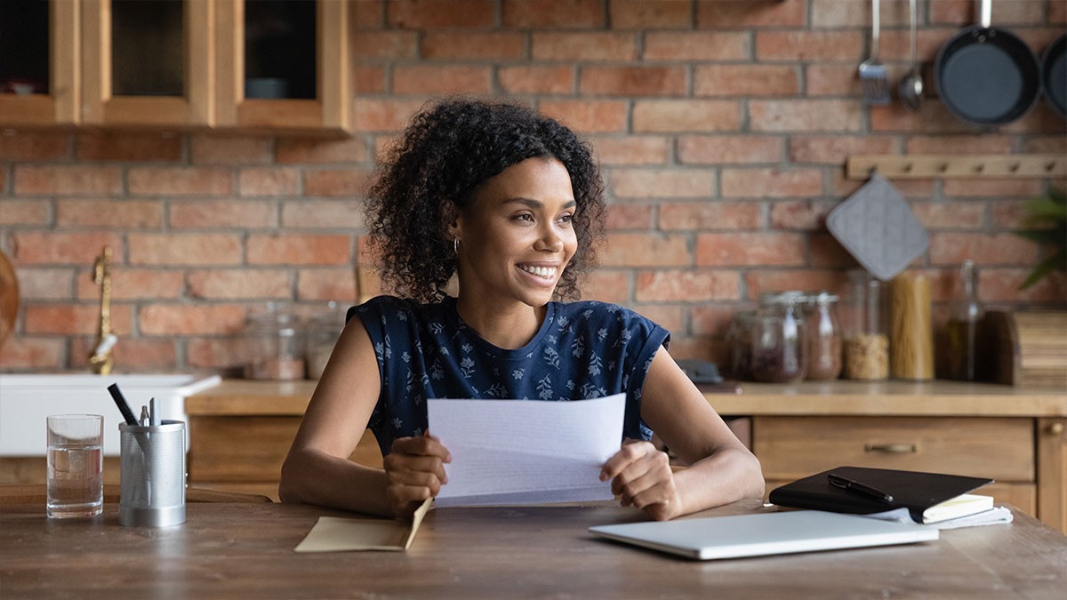 Smiling woman holding papers at a kitchen table with a laptop, against a brick wall background.