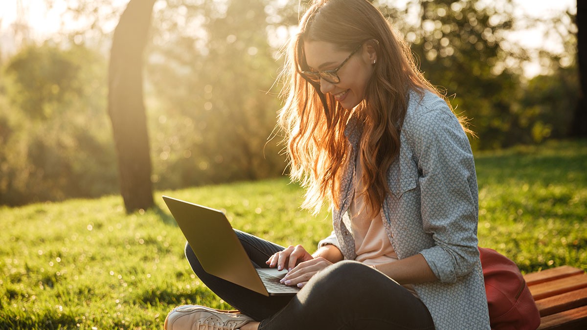 Woman sitting on grass in a park using a laptop, backlit by sunlight. She is relaxed and focused, surrounded by trees and greenery.