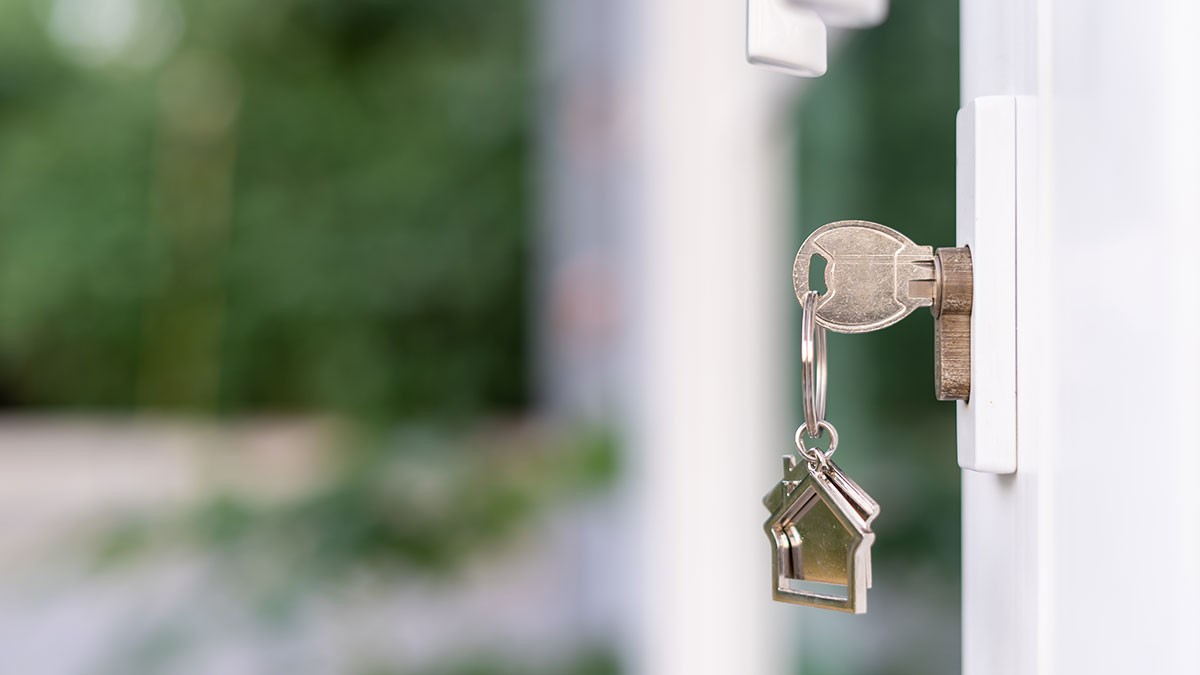 A key with a house-shaped keychain is inserted into a white door lock, with a blurred green background.