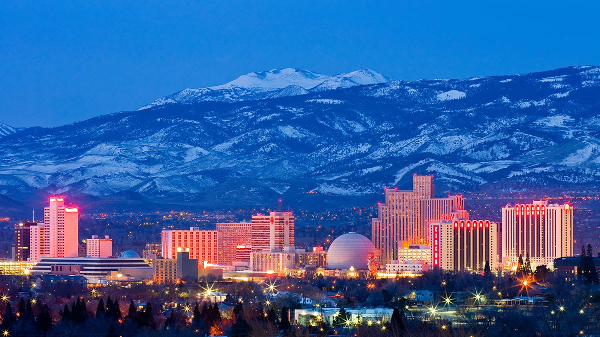City skyline at dusk with illuminated buildings and a mountainous backdrop under a clear blue sky.