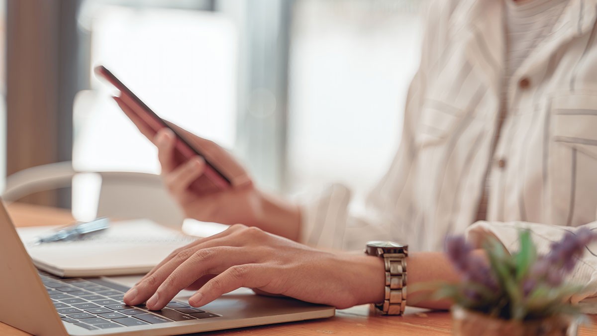 Person using a laptop and holding a smartphone at a wooden table, with a plant and notebook nearby.