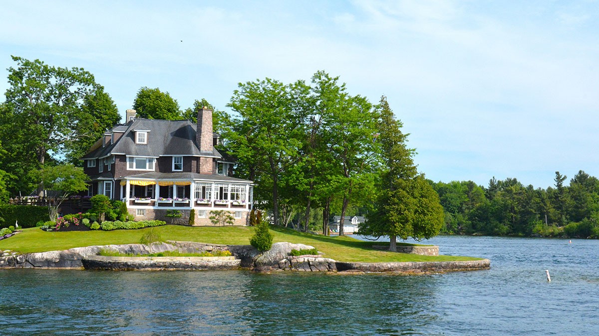 A picturesque house with a chimney on a small tree-lined island, surrounded by calm blue water under a clear sky.