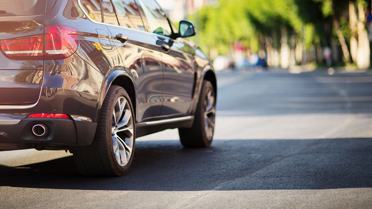 Close-up of a shiny SUV driving on a sunlit urban street with blurred green trees and buildings in the background.