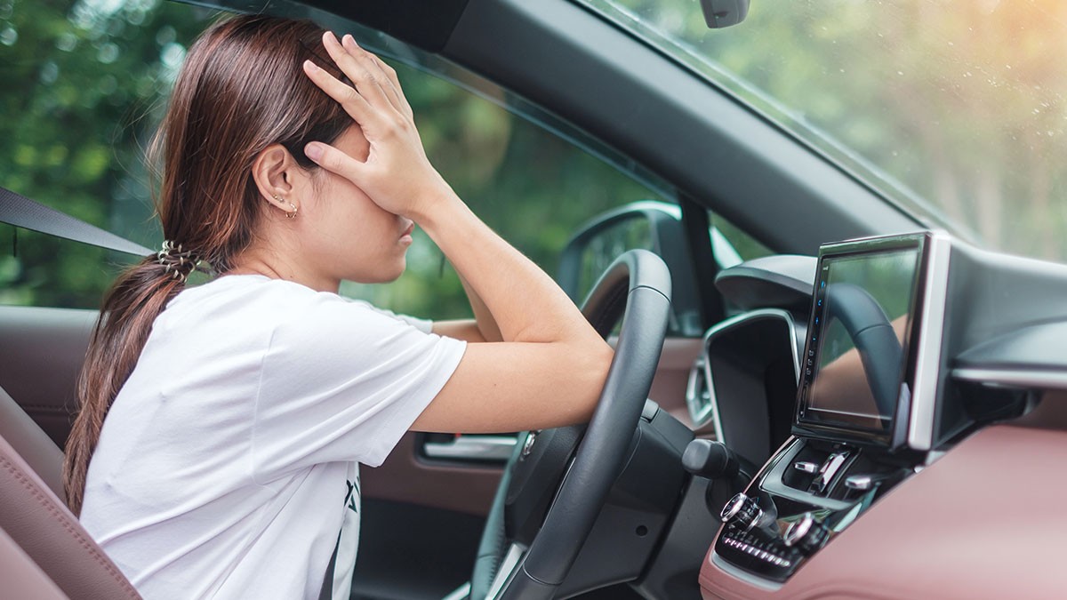 Woman sitting in a car with one hand on the steering wheel, the other covering her face, appearing stressed or frustrated.