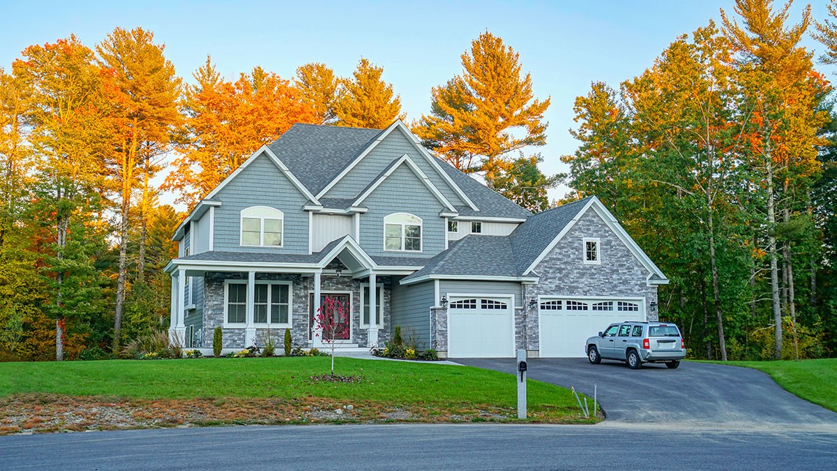 Two-story gray house with a two-car garage, surrounded by autumn trees. A car is parked in the driveway under a clear blue sky.