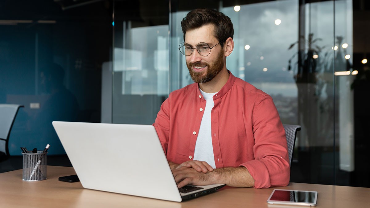 Man with a beard and glasses smiling while using a laptop in a modern office. A smartphone and pen holder are on the table.