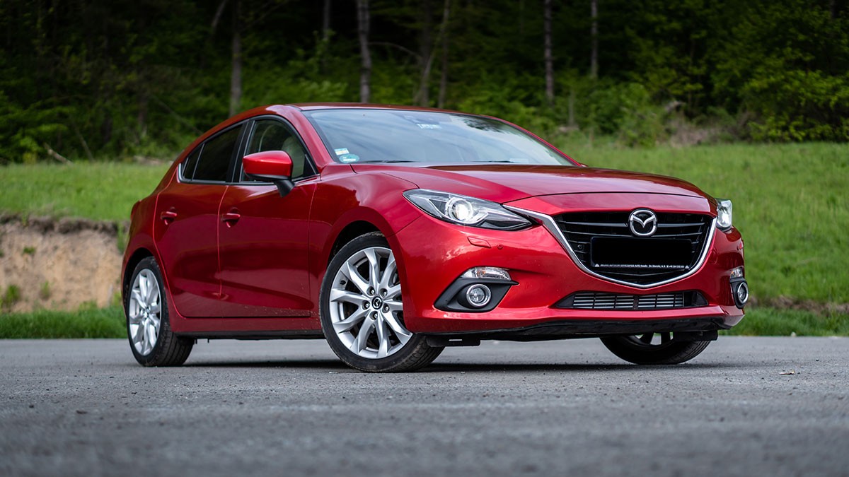 Red Mazda compact car parked on pavement with a grassy embankment and trees in the background.