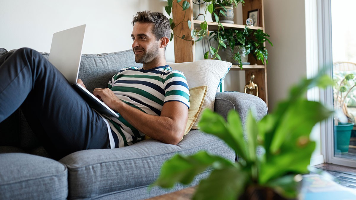 Man in striped shirt using a laptop on a gray couch in a living room with plants and a wooden shelf.