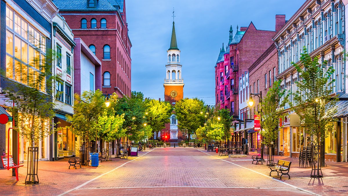 A quaint, empty street lined with shops, trees, and benches, leading to a church with a tall steeple under a twilight sky.