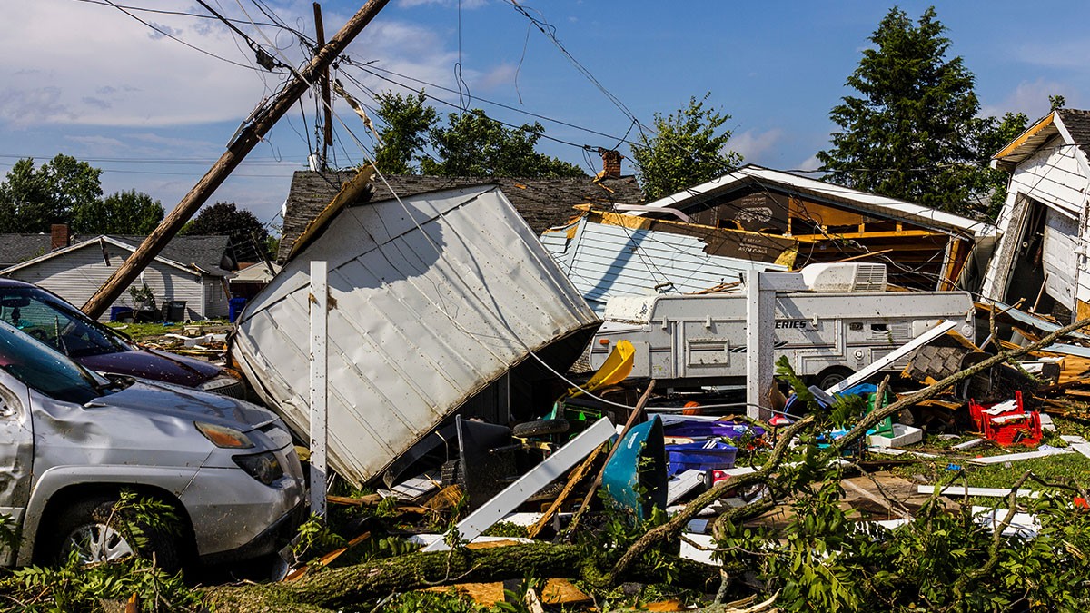 Severe storm damage showing a crushed car, fallen power lines, and destroyed buildings, with debris scattered in a residential neighborhood.