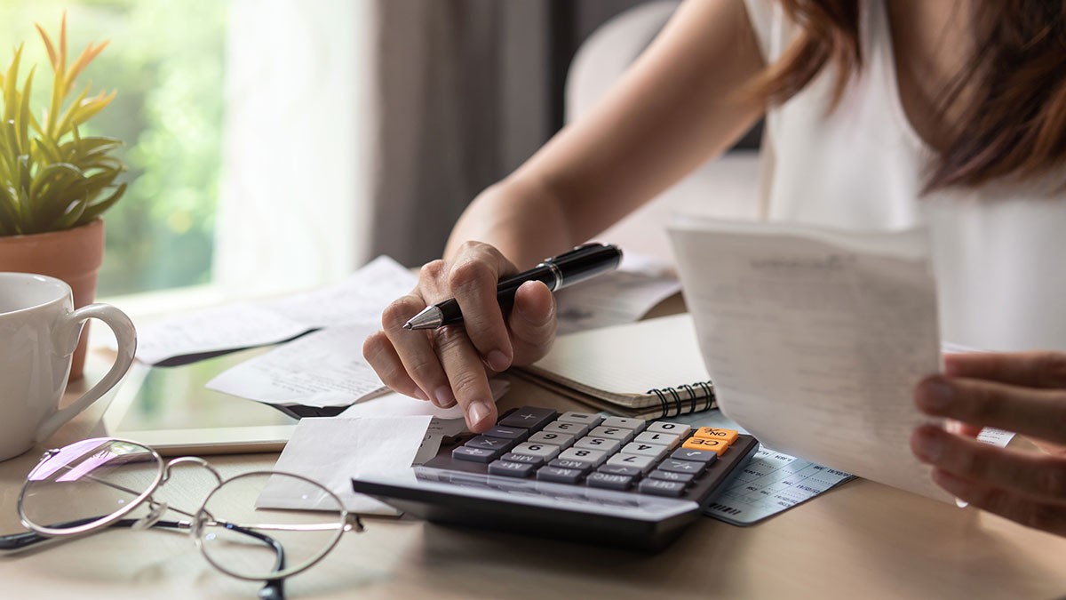 Person uses a calculator and holds a receipt at a desk, surrounded by glasses, a notebook, and a potted plant.