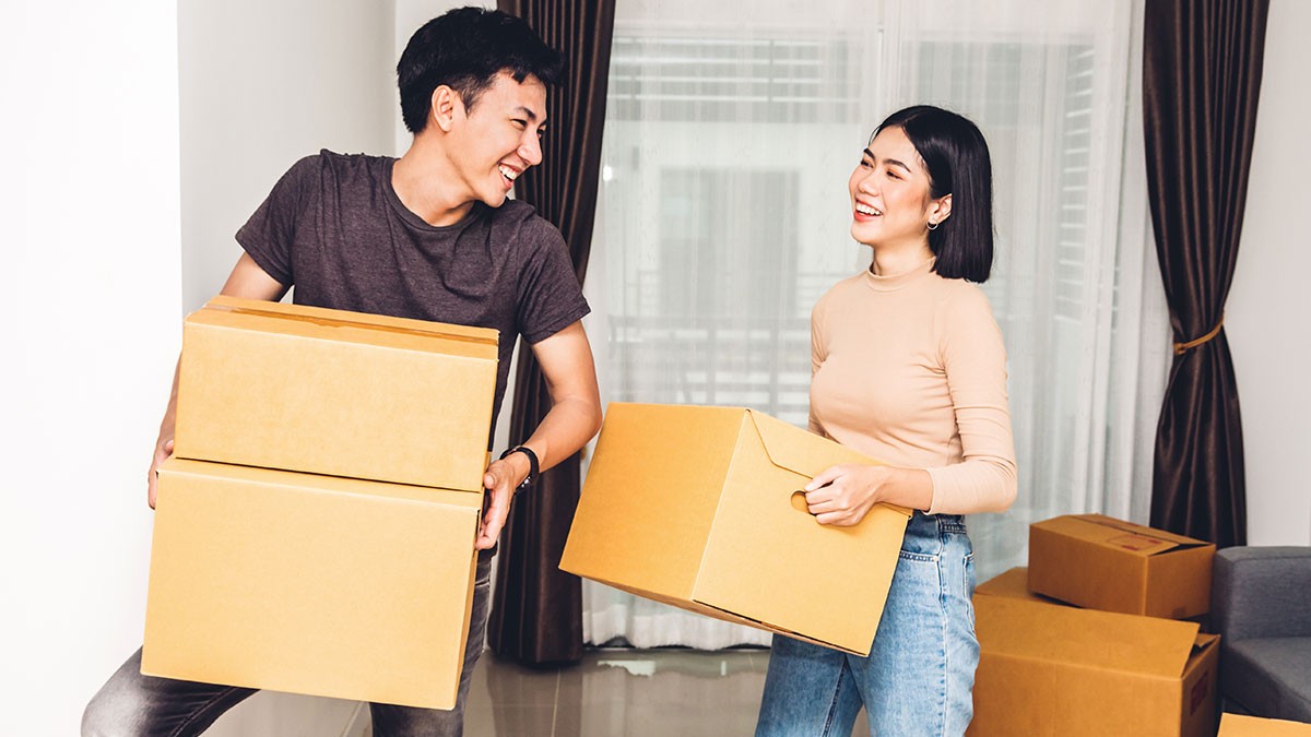 A smiling couple carries cardboard boxes in a bright room with curtains, suggesting they are moving or unpacking.