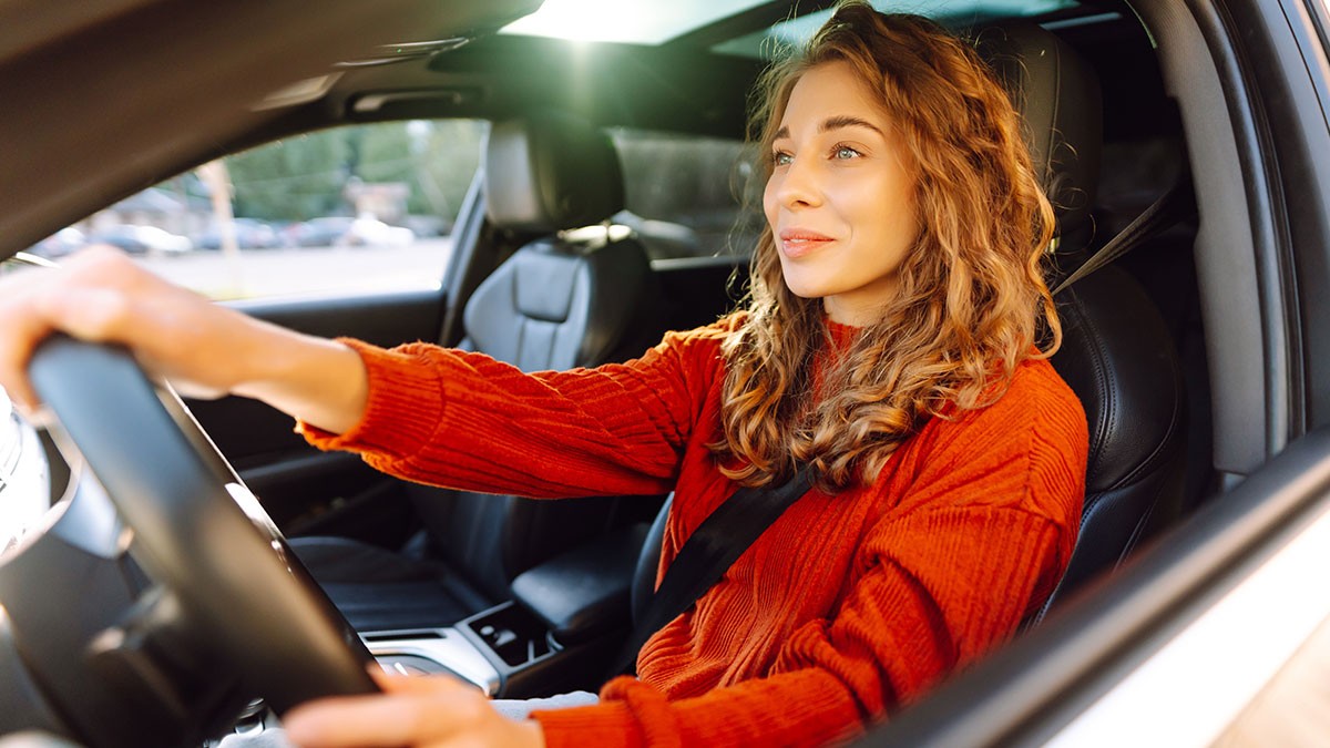 A woman with curly hair, wearing a red sweater, drives a car with a content expression. Sunlight filters through the windows.