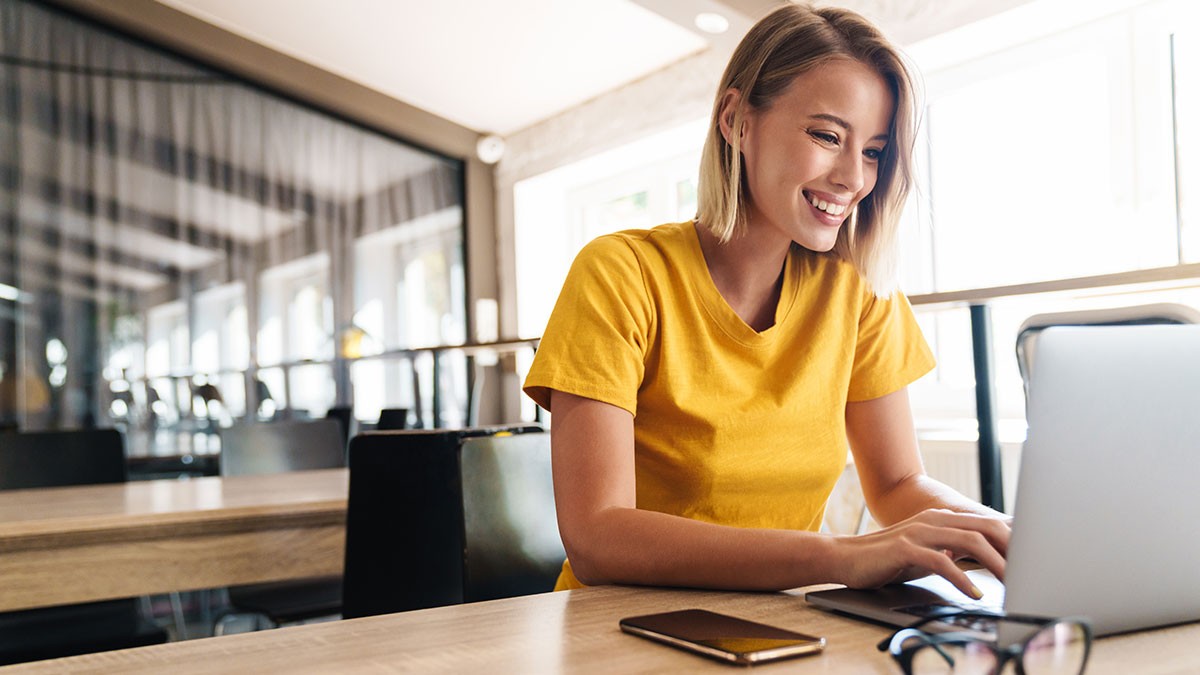 Smiling woman in a yellow shirt using a laptop at a modern cafe, with a smartphone and glasses on the table.