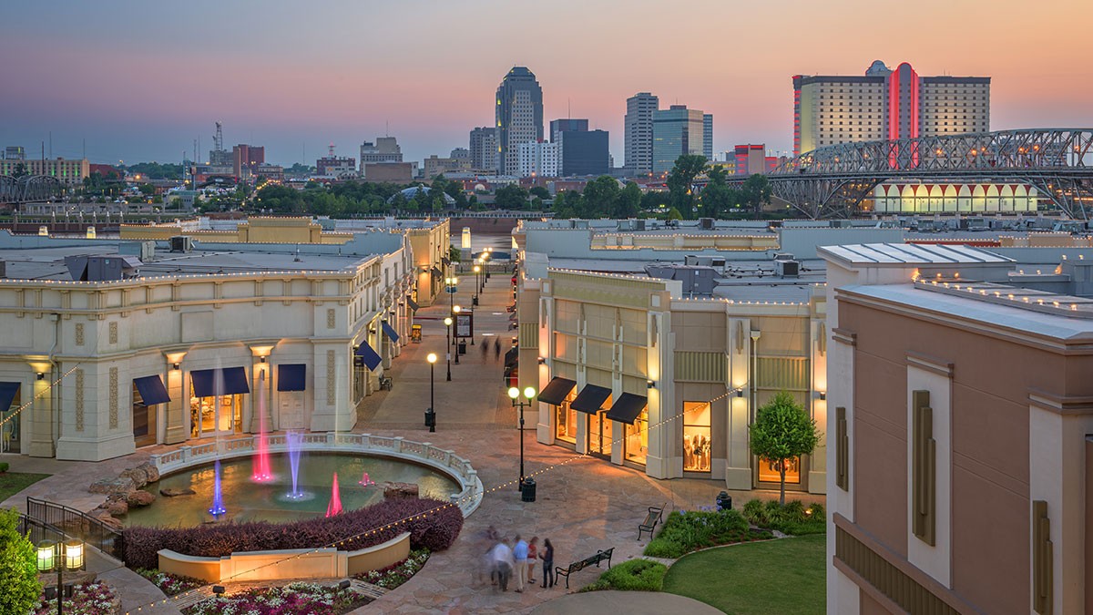 Aerial view of a shopping district at sunset, featuring fountains, streetlights, and buildings, with a city skyline and bridge in the background.