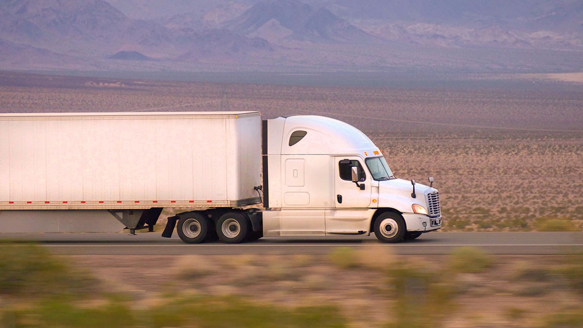 White semi-truck with enclosed trailer driving along a desert highway with distant mountains under a hazy sky.