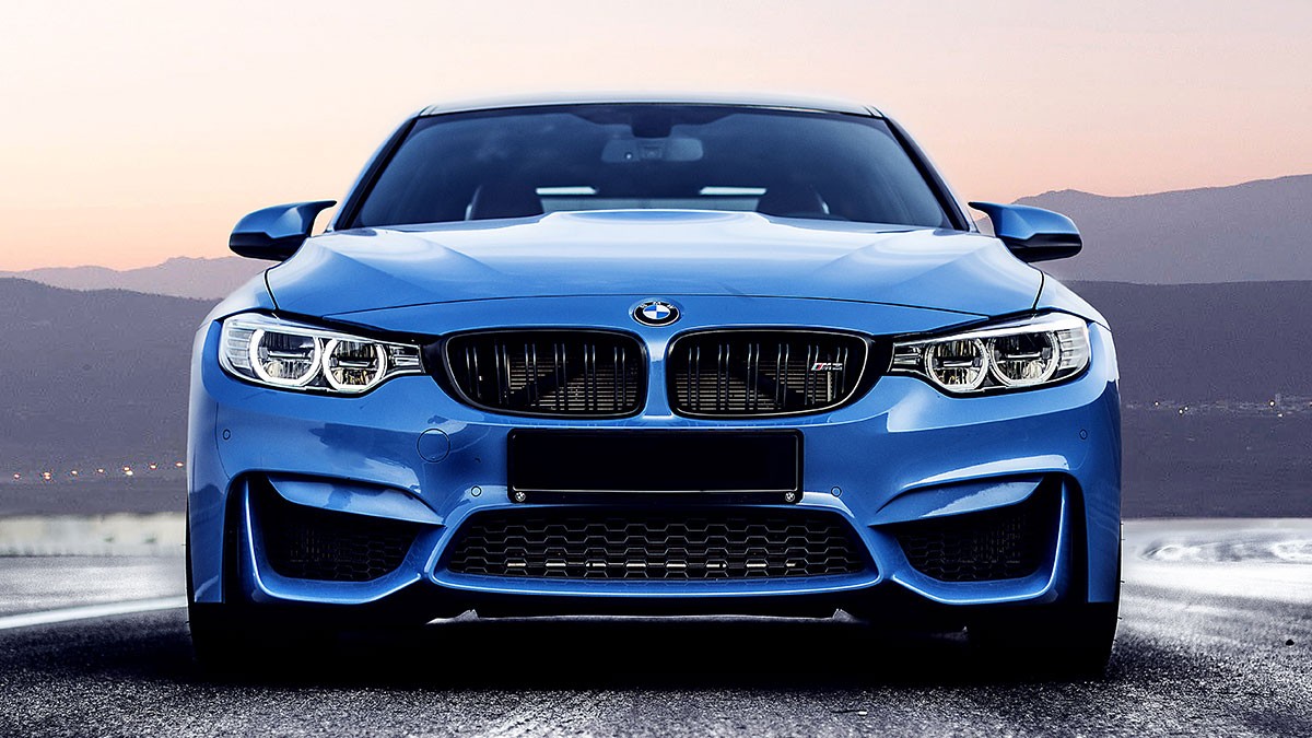 Front view of a blue BMW car on a road with a mountainous backdrop at dusk. The car's headlights are on, and the sky is a gradient of pink and blue.