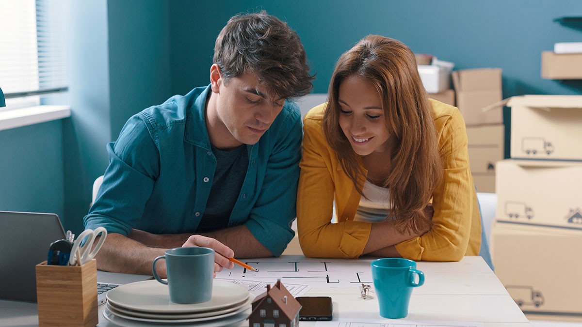 A couple reviews a floor plan together at a table, surrounded by moving boxes, with mugs and a model house nearby.