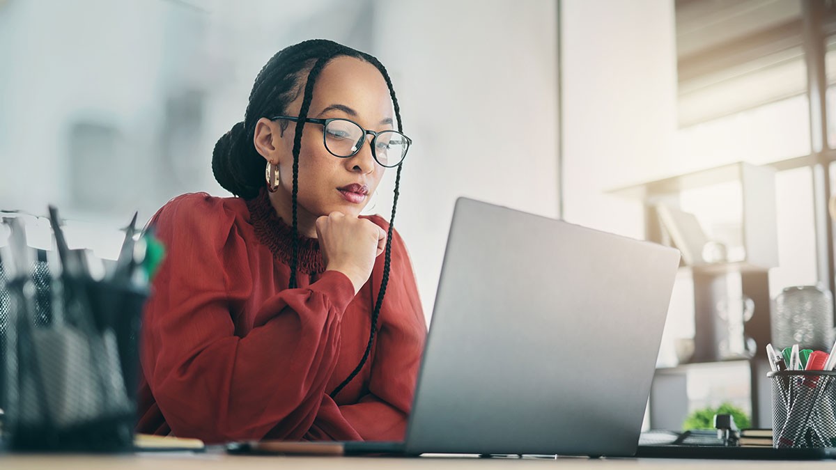 A woman in a red blouse and glasses works intently on a laptop at a desk, with office supplies and soft lighting in the background.