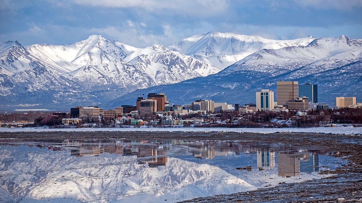 City skyline with tall buildings against snow-capped mountains, reflected in a calm body of water under a cloudy sky.