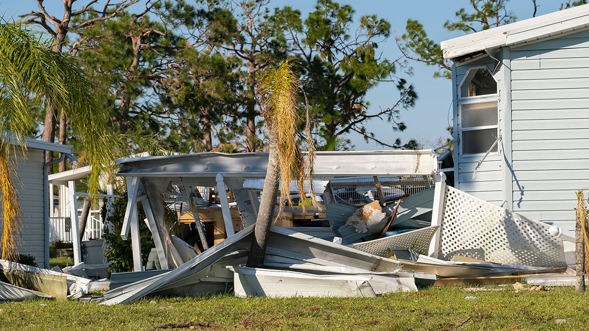Damaged mobile home with collapsed porch, debris, and fallen palm trees, set against a background of green trees under a clear blue sky.