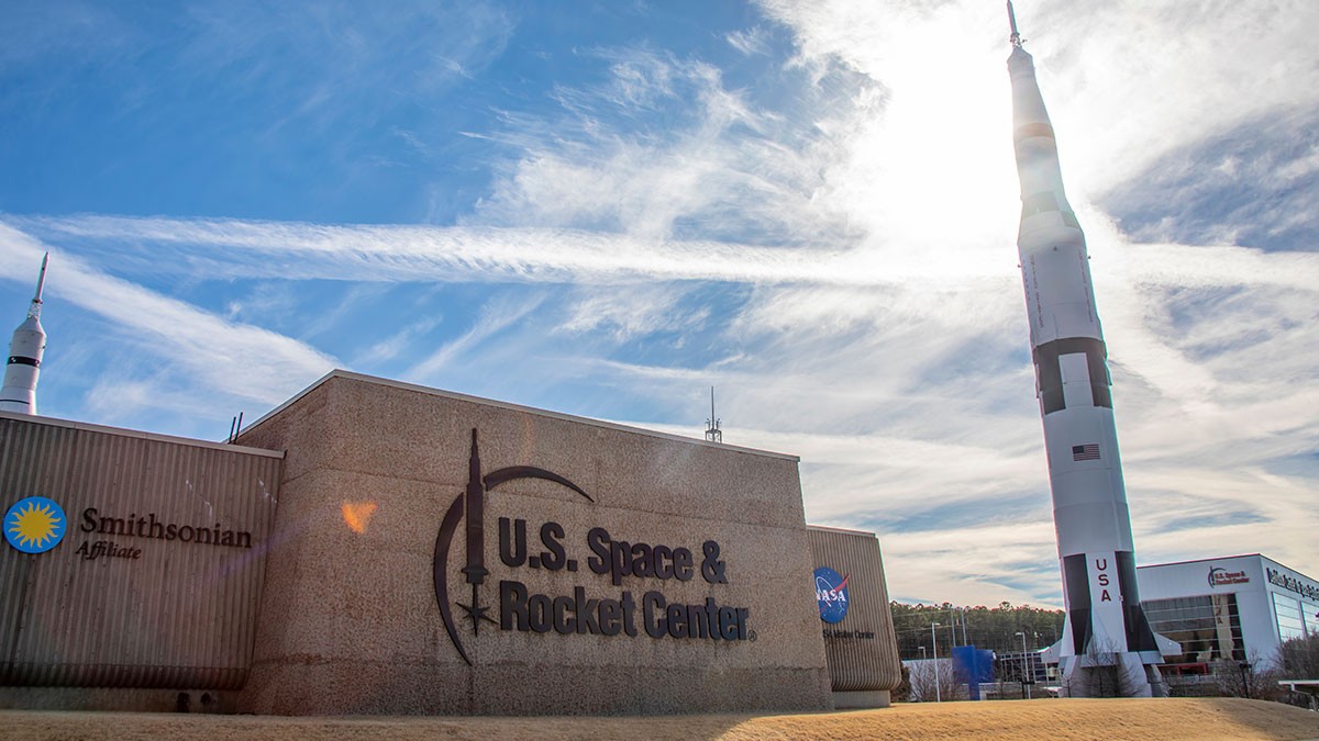 U.S. Space & Rocket Center building and sign with a tall rocket on display under a bright blue sky with wispy clouds.