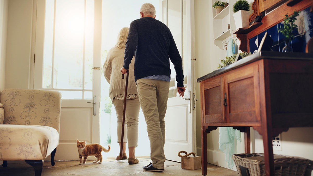 An elderly couple walking with a cane towards an open door, followed by a small orange cat in a cozy, well-decorated home.