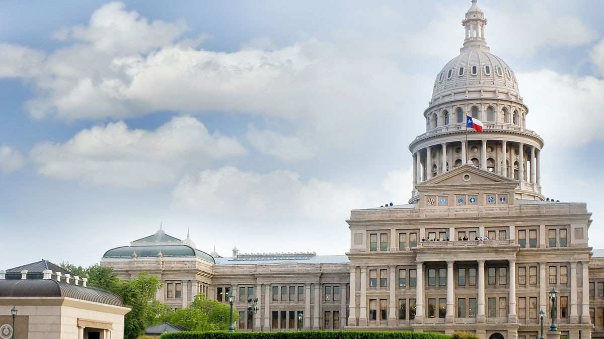 The Texas State Capitol building with its dome and a Texas flag, set against a cloudy sky.