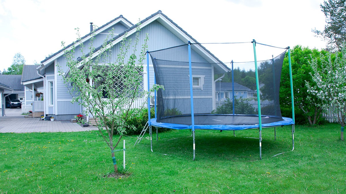 A large trampoline with a safety net stands on the green lawn in front of a gray house in a suburban setting.