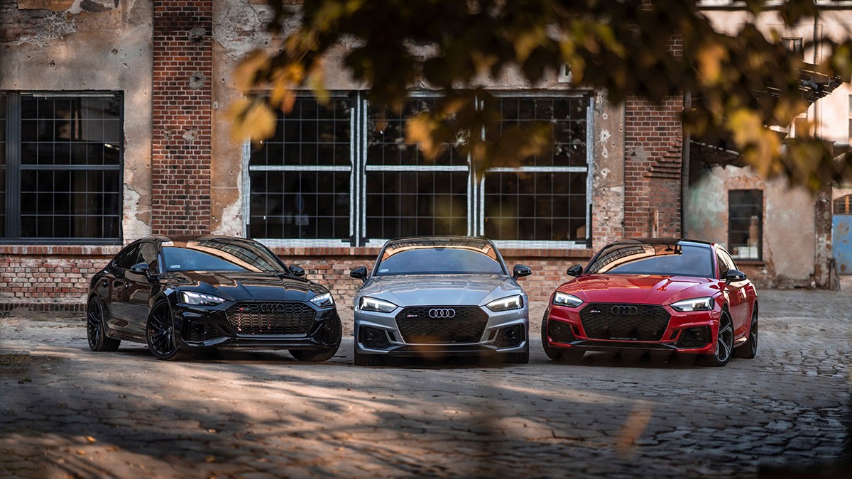 Three sleek cars, black, gray, and red, parked side by side on a cobblestone street, with an industrial brick building in the background.
