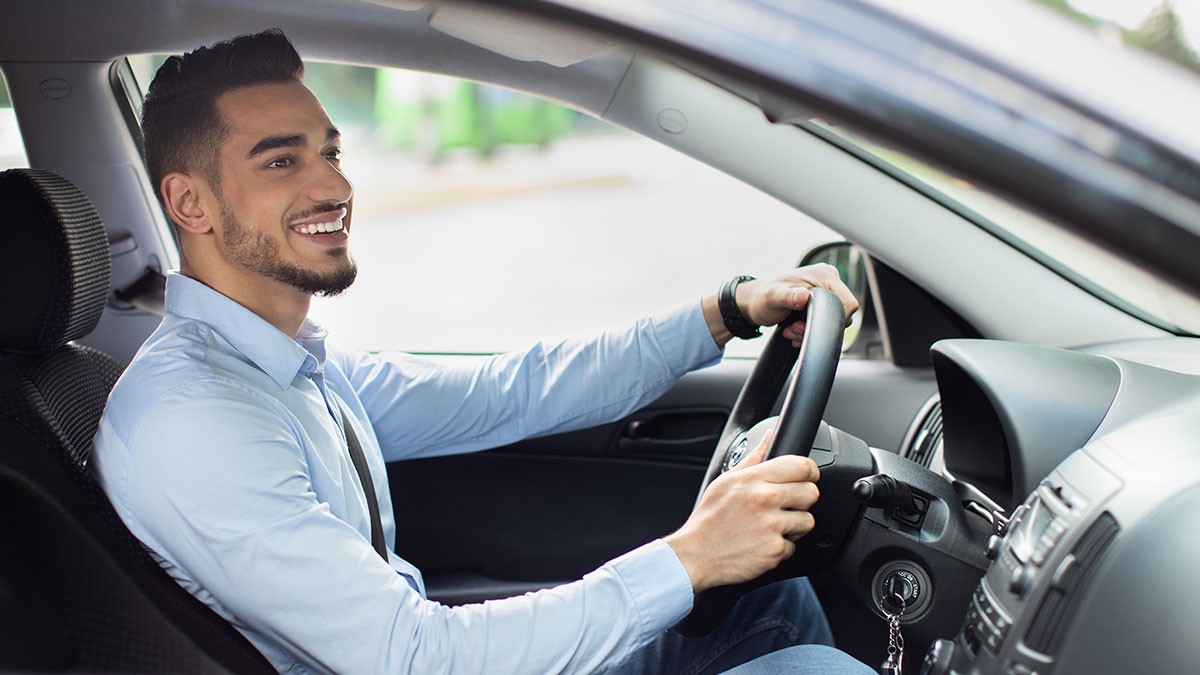 Man smiling while driving a car, wearing a light blue shirt, with hands on the steering wheel, and a blurred background outside the window.