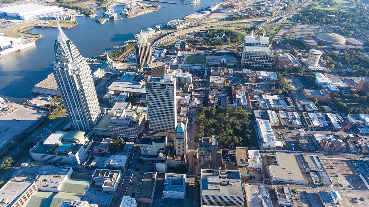 Aerial view of a cityscape with tall buildings, a river, and wide roads. The skyline is dominated by a pointed skyscraper near the water.
