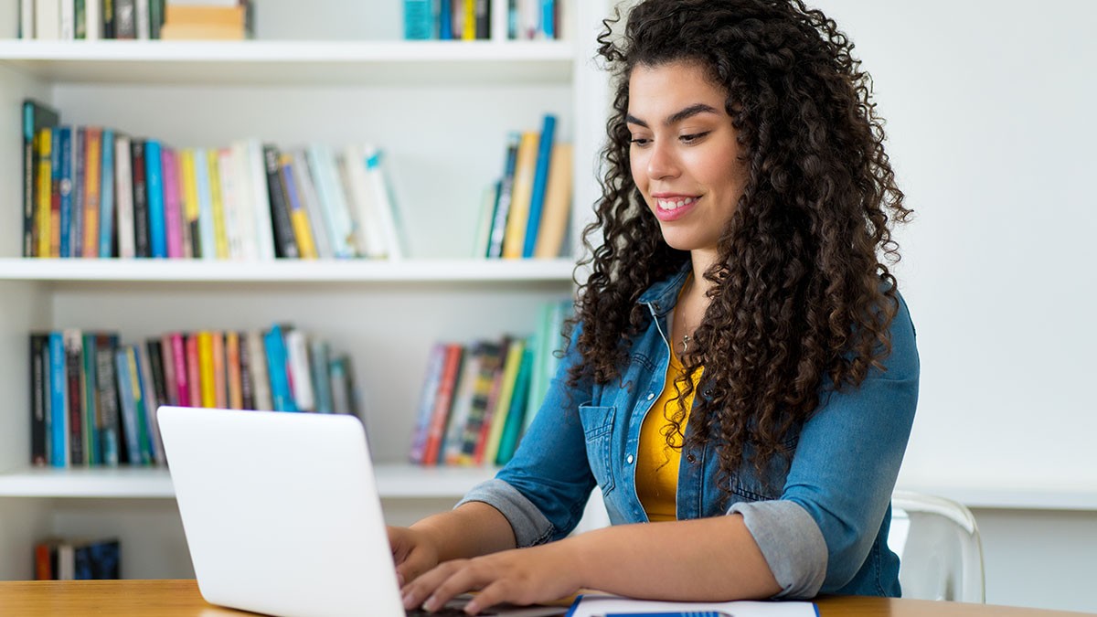Young woman with curly hair wearing earbuds, smiling while working on a laptop at a desk with a bookshelf behind her.