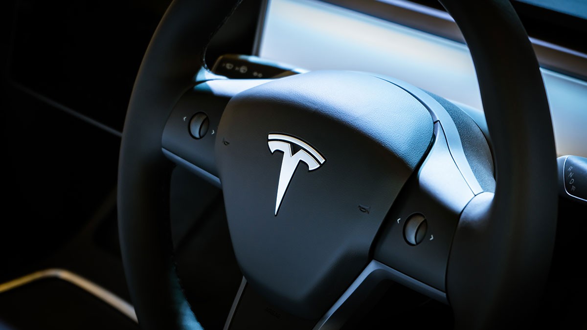 Close-up of a Tesla steering wheel, showcasing the Tesla logo prominently in the center, with a sleek and modern dashboard in the background.