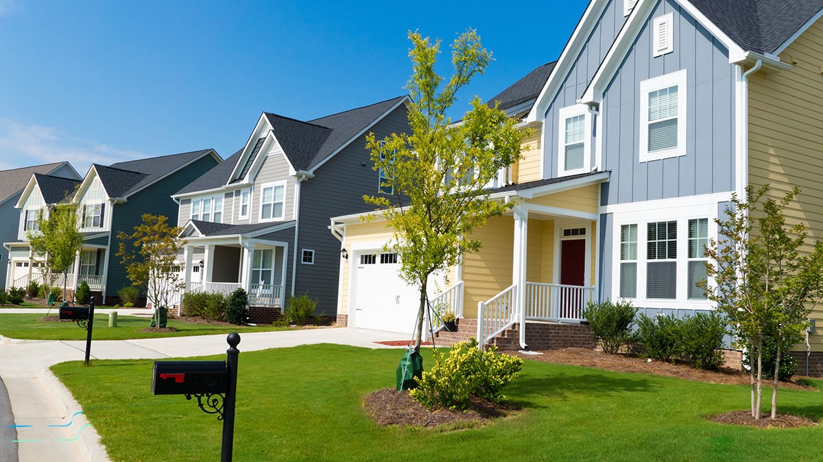 A row of modern suburban houses with manicured lawns, trees, and mailboxes under a clear blue sky.